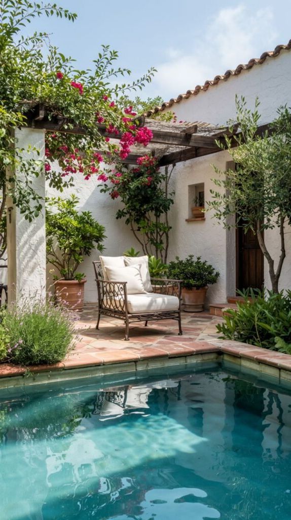 A small pool is located next to a patio area with terra cotta colored tiles. An outdoor armchair with a white cushion is placed under a wooden pergola covered in flowering vines with pink blooms. Potted plants are arranged around the chair and near a white stucco wall.