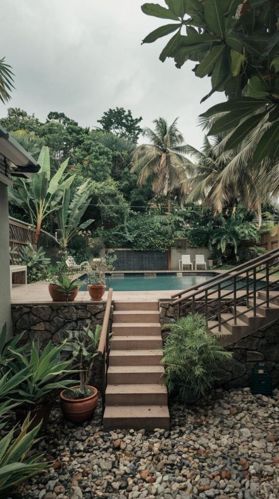 A multi-level pool area is accessed by stone steps leading down from a stone-faced wall. The pool itself is rectangular and surrounded by lush, tropical plants including large-leaved varieties and banana trees. Outdoor chairs are visible by the pool.